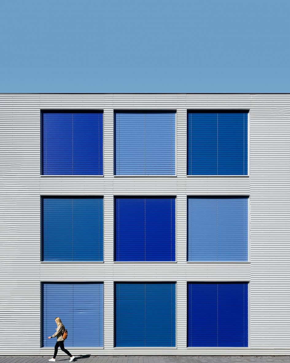 Grey building with 9 square windows in various shades of blue. A person is walking past the building.